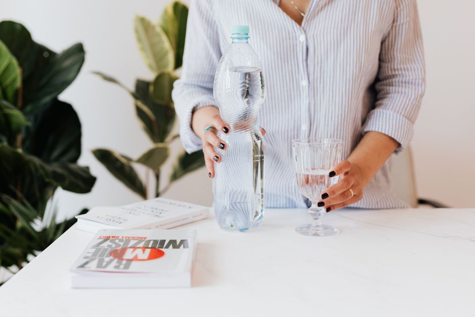 Unrecognizable female in shirt with stripes standing near white table and holding bottle and glass of water in hands